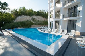 A swimming pool with lounge chairs, a building with balconies, and a hillside in the background.