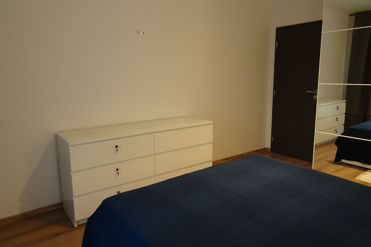 A bedroom with a bed, a white dresser, a dark-colored wardrobe, and a mirror.