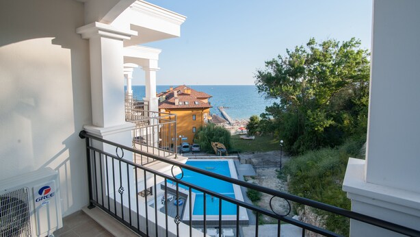 Un balcone con vista su una piscina, un edificio con tetto rosso e il mare sullo sfondo.