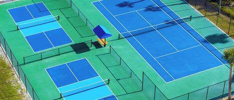 An aerial view of a tennis court complex with multiple courts, surrounded by a fence and palm trees.