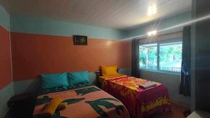Habitación con dos camas, una con colcha estampada azul y verde, y la otra con estampado rojo y amarillo. Se ve vegetación a través de la ventana.