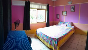 Una habitación con una cama, una silla, un ventanal con cortinas, un ventilador y un cuadro en la pared.