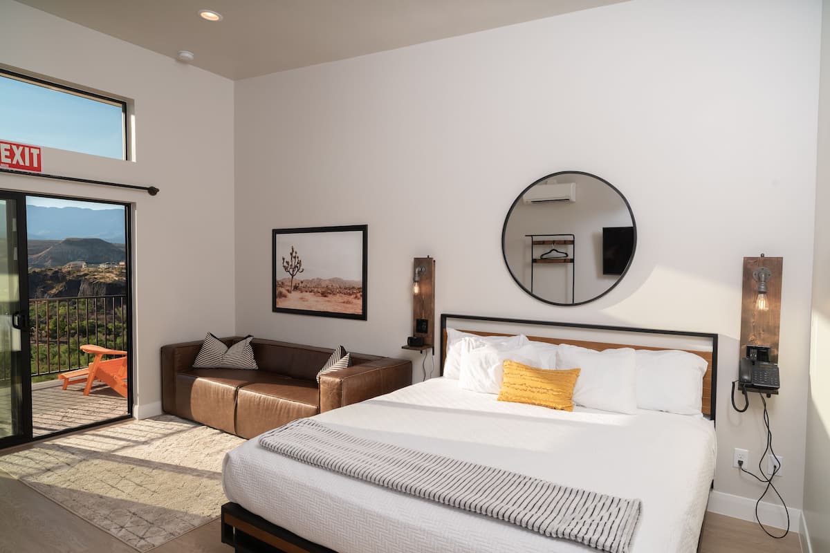 A modern hotel room with a large bed, a sofa, a balcony with an exit sign, a framed picture, a round mirror, and a telephone on the wall.