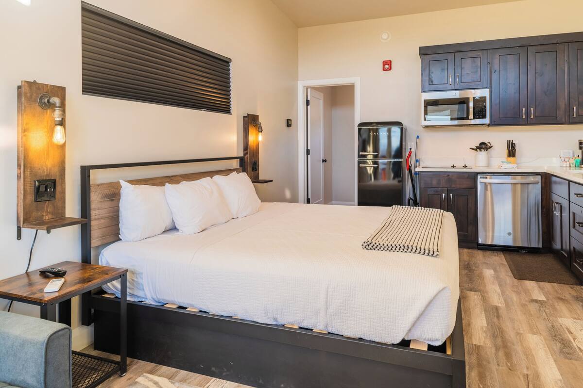 A modern hotel room with a large bed, a small desk, a built-in wardrobe, and a kitchenette with a microwave and refrigerator.