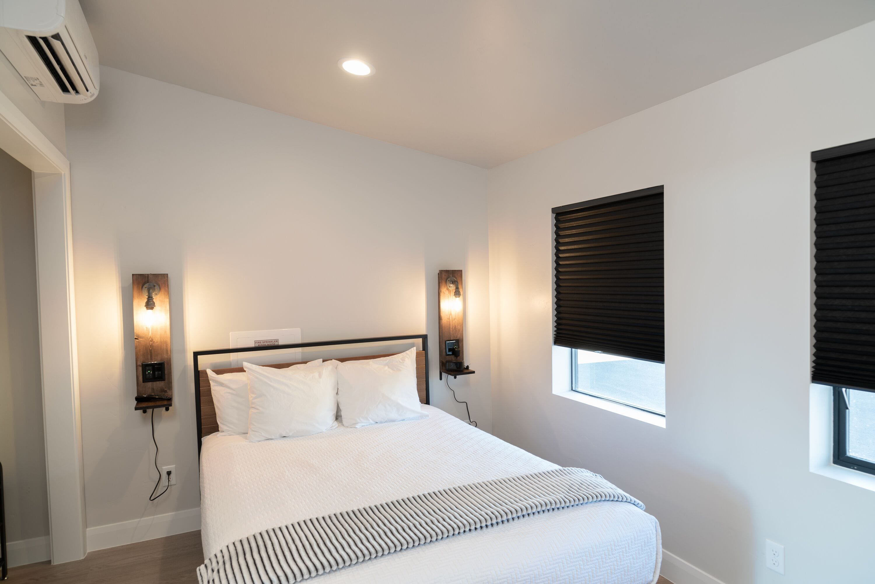 Une chambre bien rangée, avec un grand lit, deux lampes fixées au mur, une fenêtre avec des stores et un appareil de climatisation.