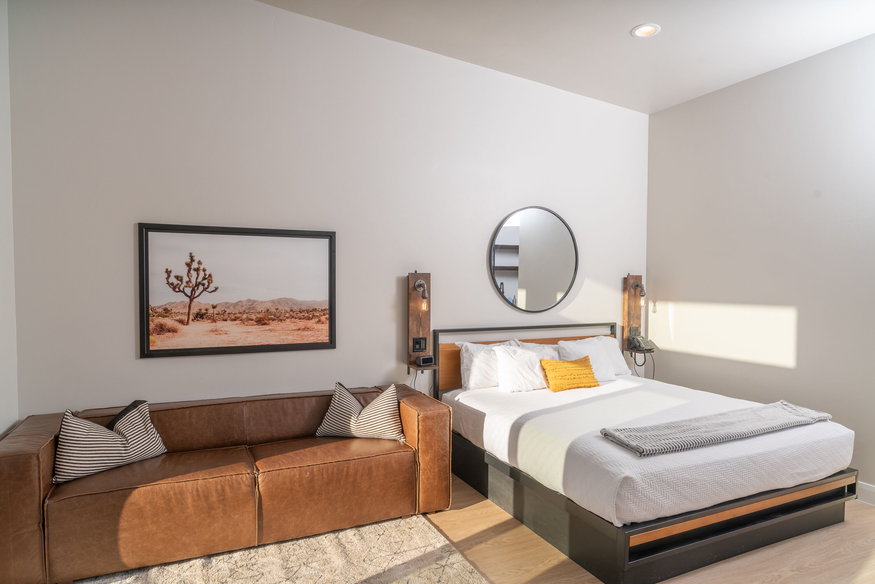 A modern bedroom with a bed, a brown leather sofa, a framed desert landscape picture, a round mirror, and a bedside table with a lamp.