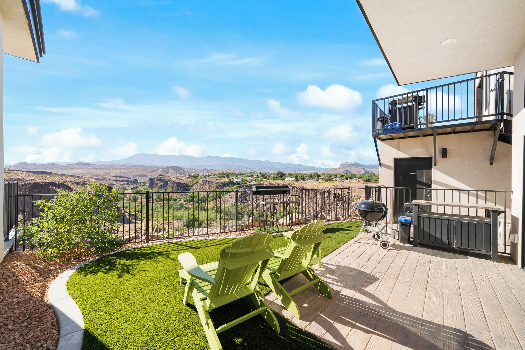 A balcony with two green Adirondack chairs, a barbecue grill, and a view of distant mountains.