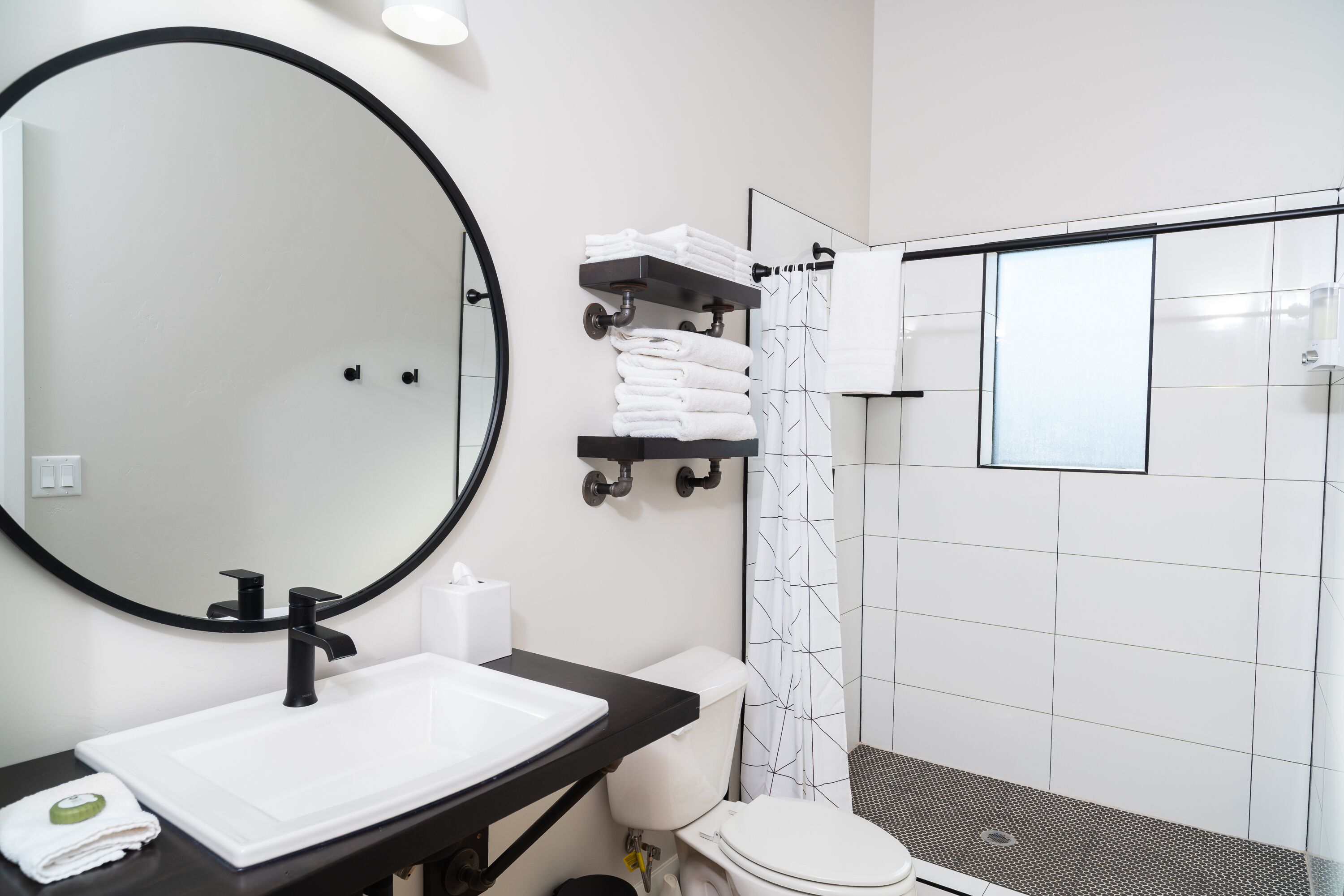 A modern bathroom with a large round mirror, a white sink, a black countertop, a toilet, and a shower area with a glass door.