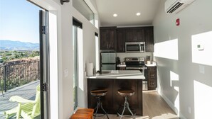 A modern kitchen with dark wooden cabinets, a stainless steel refrigerator, and a microwave. There are two stools and a small table. - The Dwellings (La Verkin)