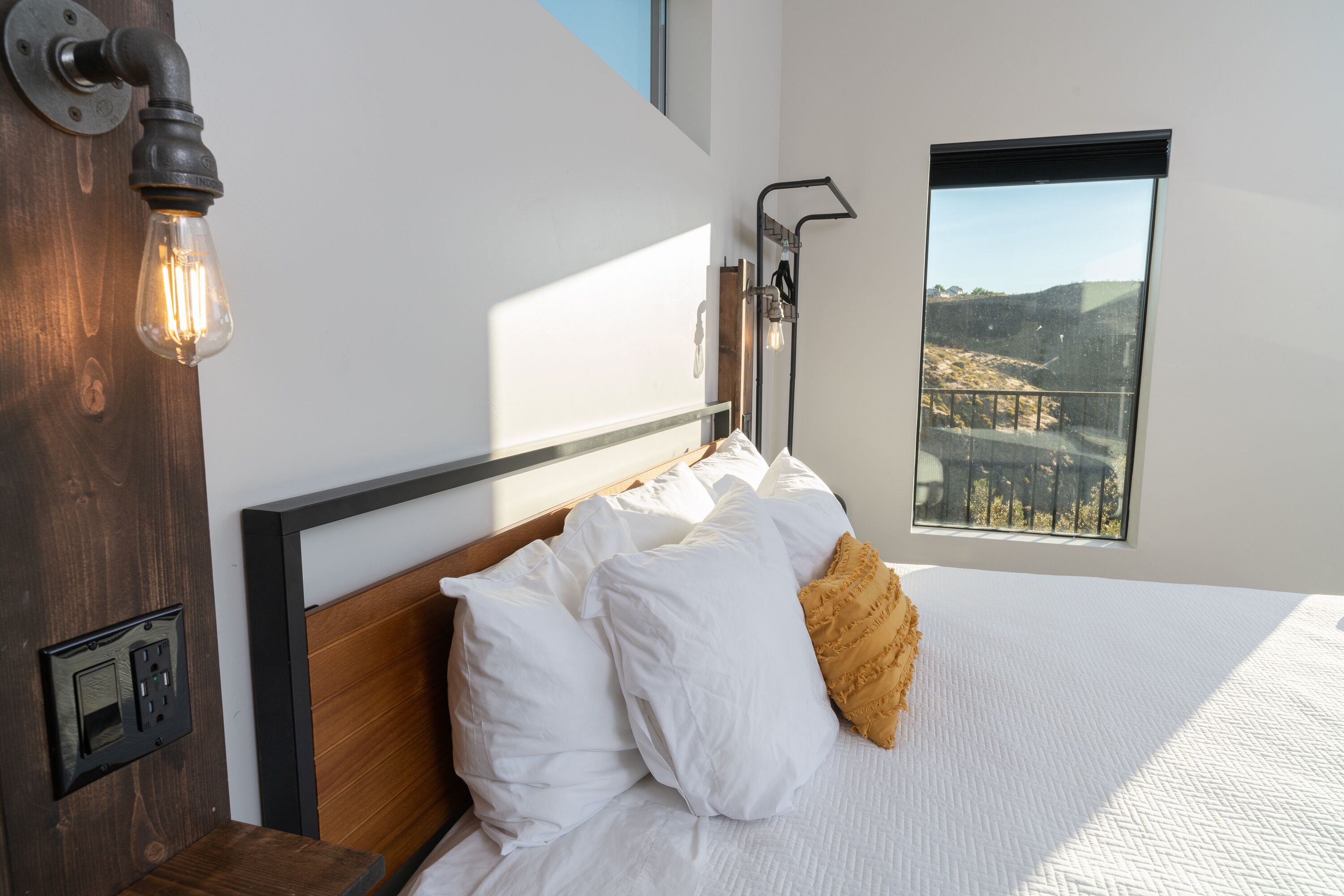 A modern bedroom with a wooden headboard, white bedding, a single yellow pillow, and a view of a landscape through a window.