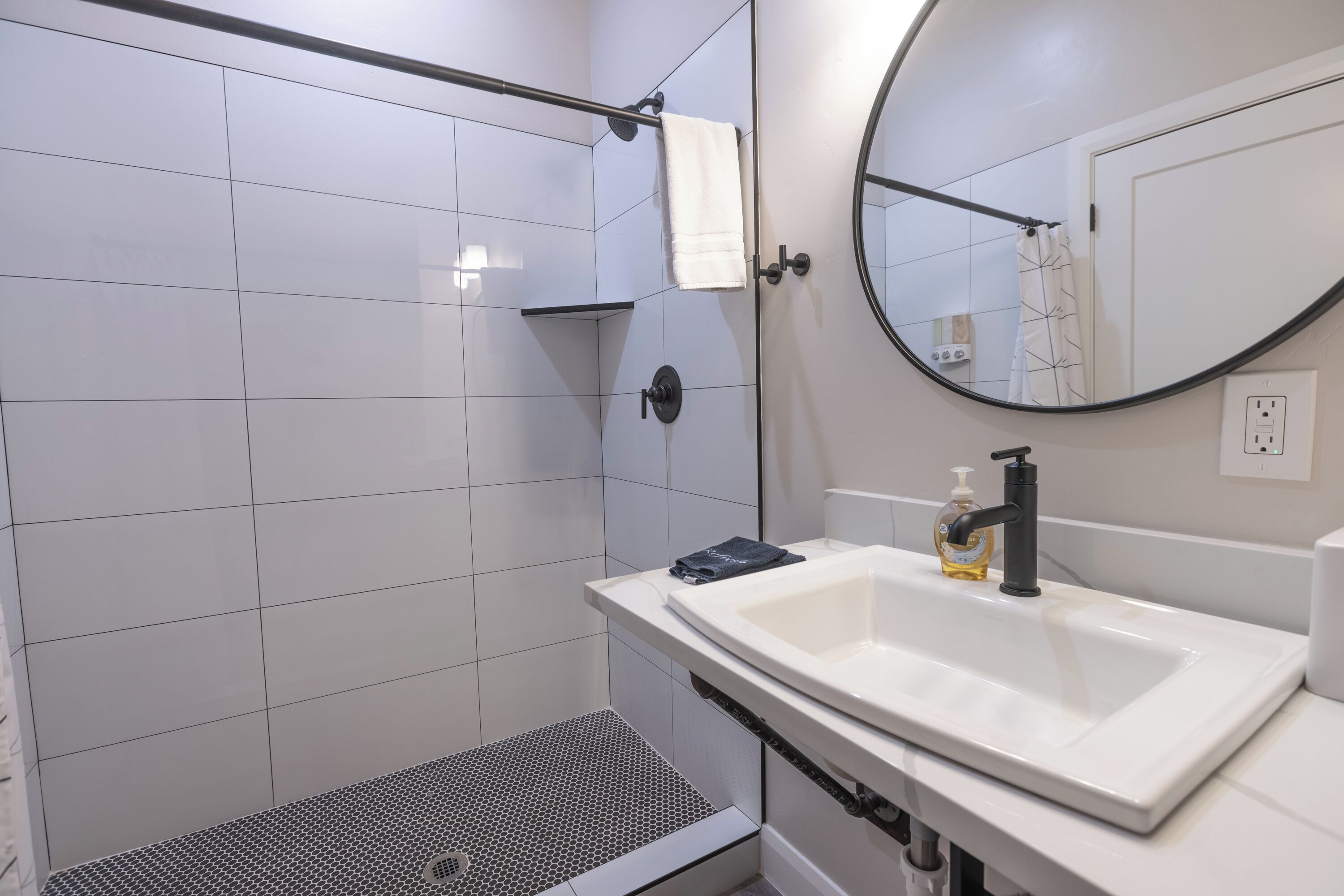 A modern bathroom with a white sink, a round mirror, and a shower area with white tiles.