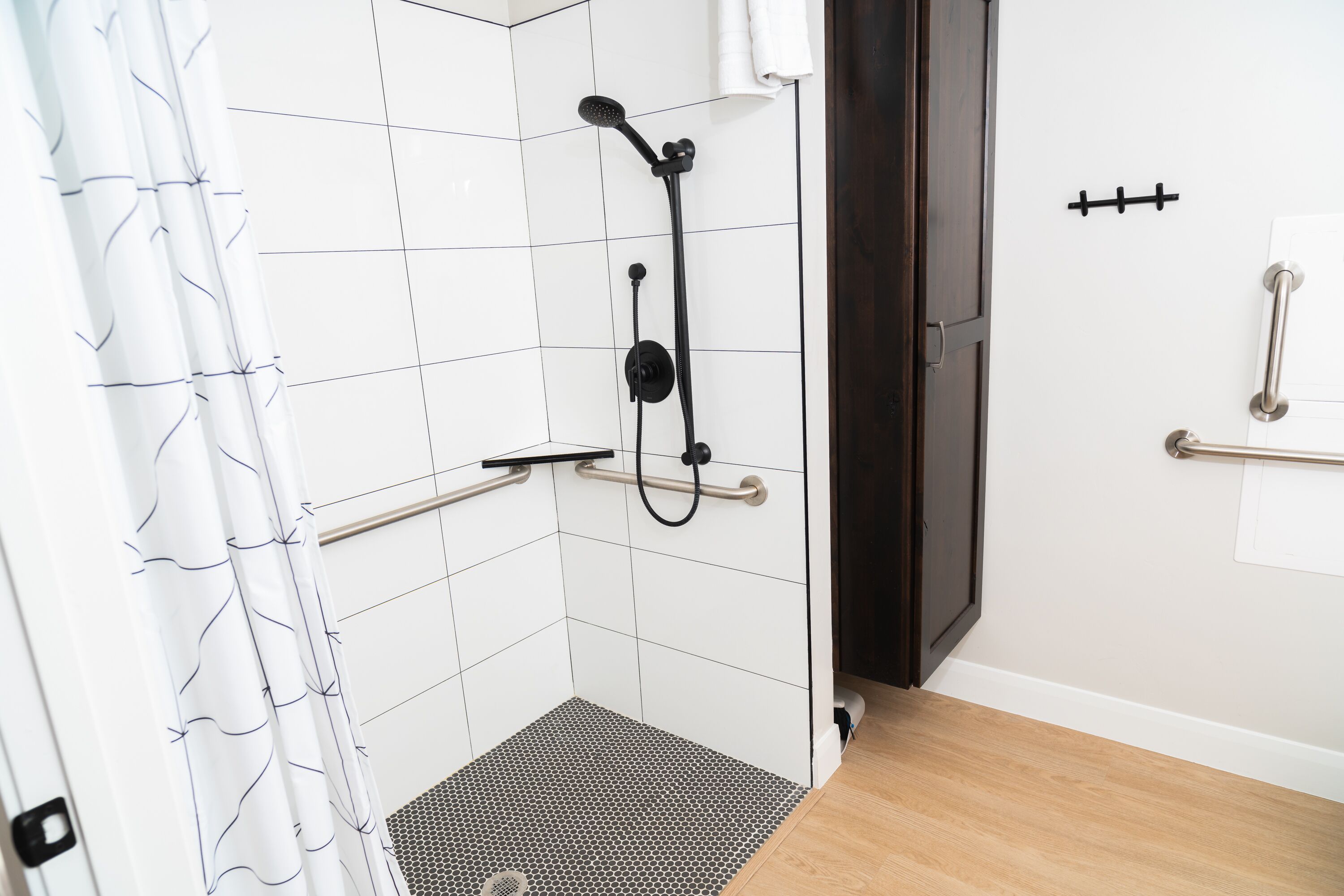 A bathroom with white tiled walls, a shower with a handheld showerhead, a grab bar, and a wooden floor.