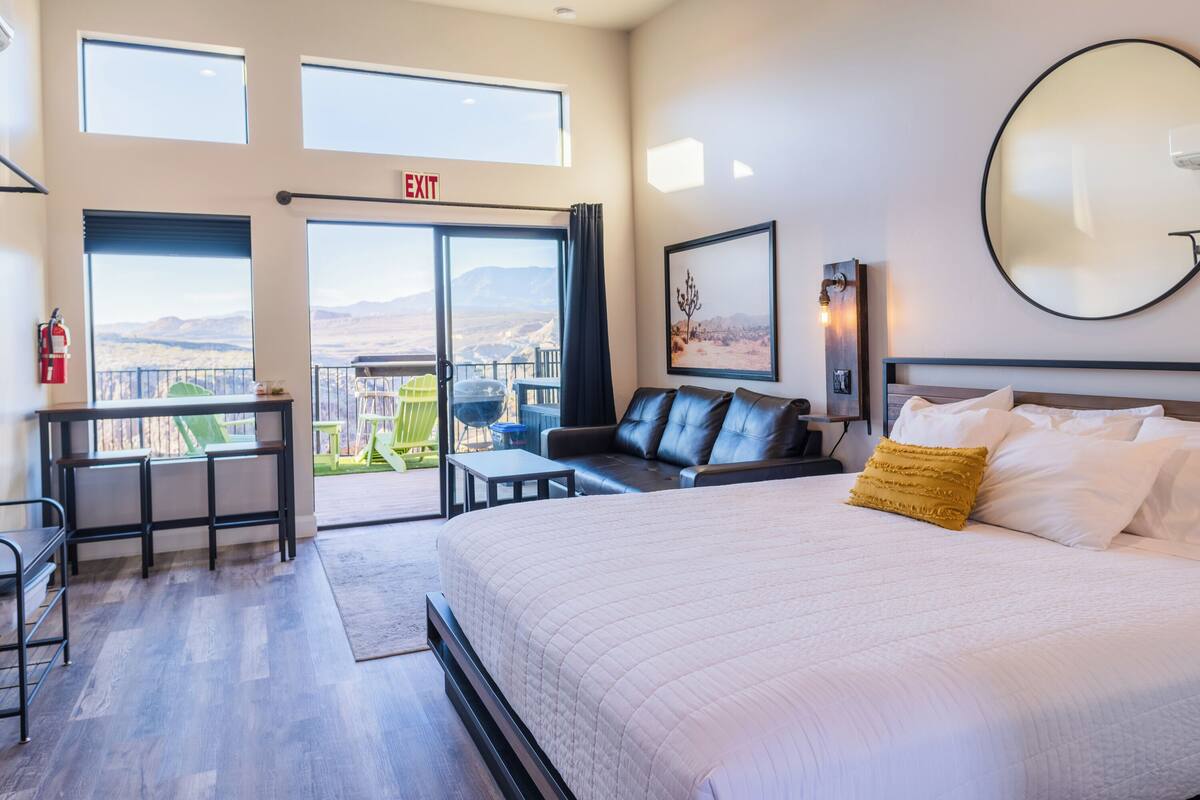 A hotel room with a large bed, a desk, a chair, and a balcony with a view of the mountains.