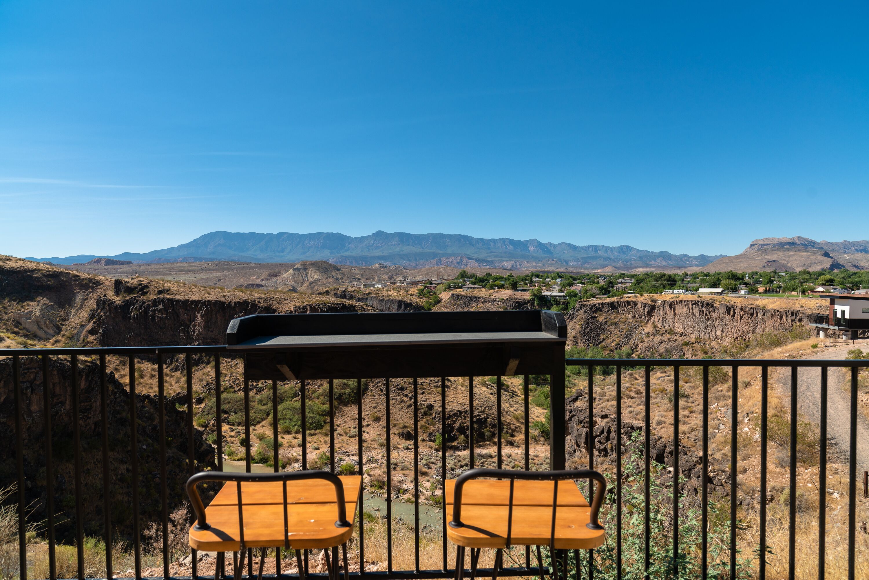 A balcony with two wooden chairs and a grill, overlooking a mountainous landscape with sparse vegetation and a few buildings in the distance.