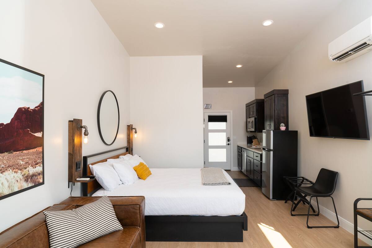 A modern hotel room with a large bed, a desk, a kitchenette, and a TV mounted on the wall.