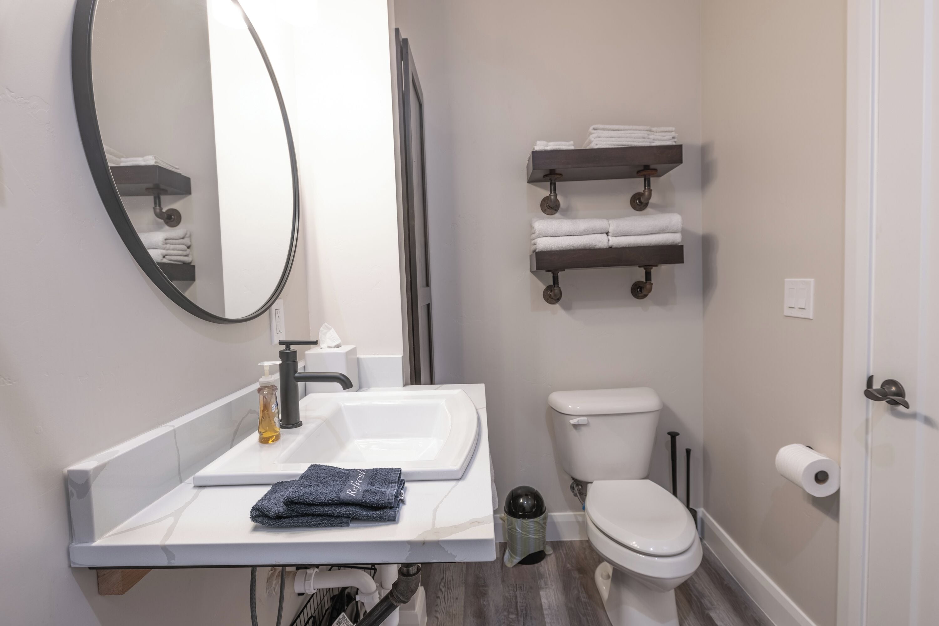 Une salle de bain moderne avec un lavabo, un miroir et des toilettes blancs. On trouve des porte-serviettes au-dessus des toilettes et une poubelle à côté du lavabo.