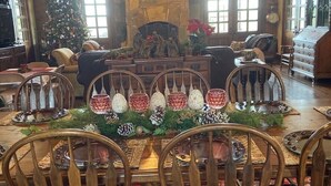 Un coin repas chaleureux avec une table en bois dressée pour six personnes, entourée de chaises en bois. Un lustre est suspendu au plafond, et un sapin de Noël est visible au fond.