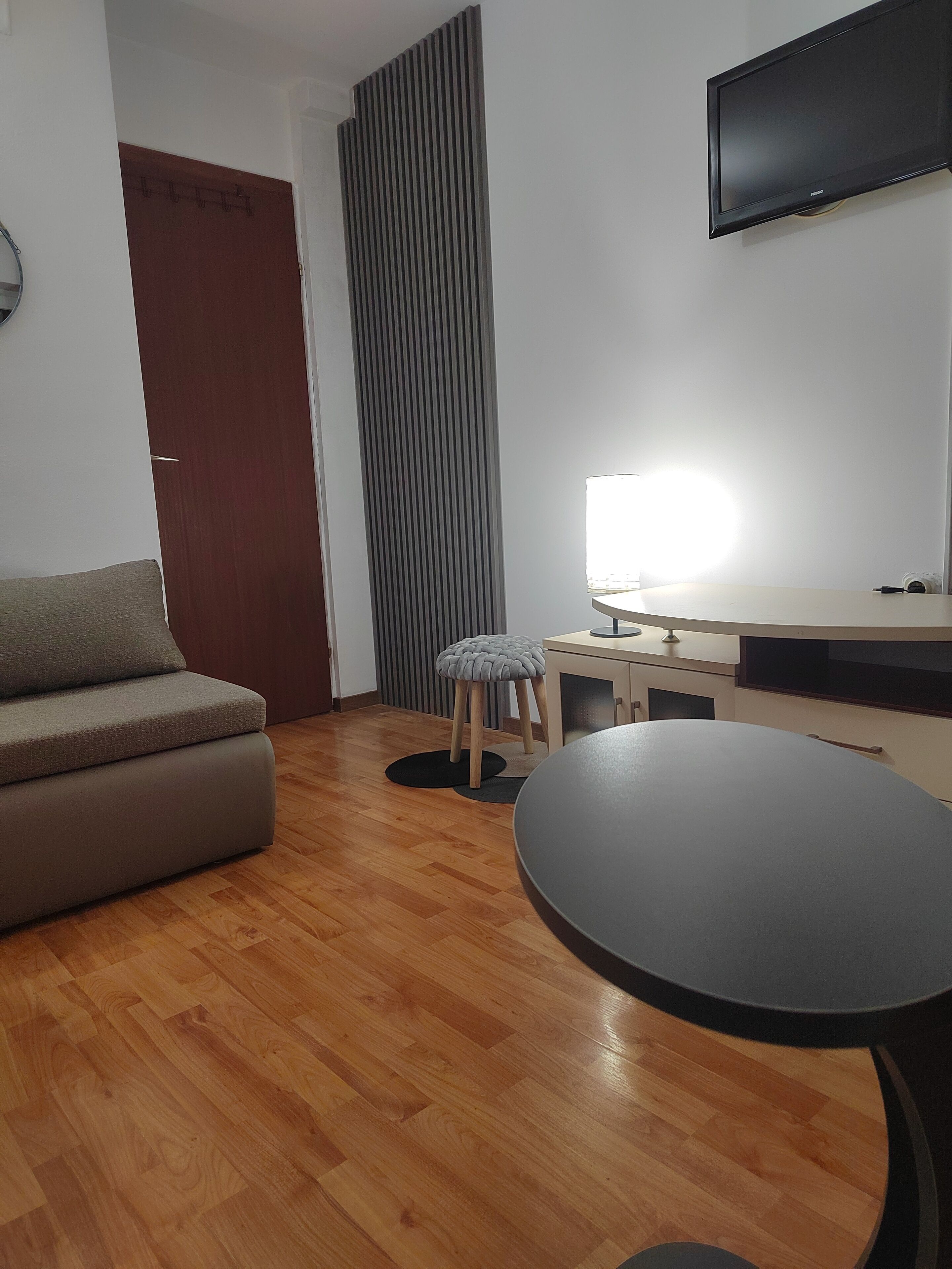 A modern living room with a sofa, a round table, a stool, a TV mounted on the wall, and a wooden floor.