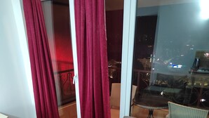 A living room with a grey sofa, a small round table with a green object, red curtains, and a view of a cityscape through a large window. - Villa Old Town (Ohrid)