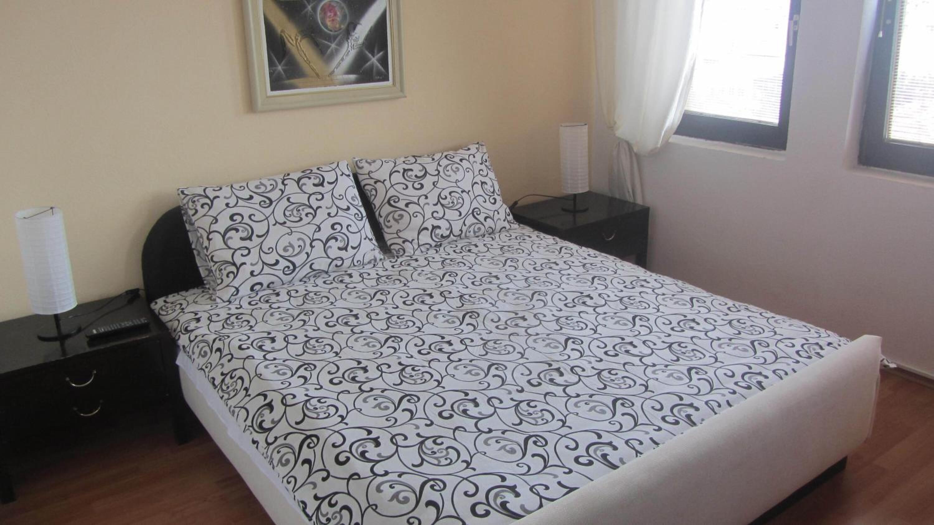 A bedroom with a patterned bedspread, a nightstand with a lamp, a framed picture on the wall, and a window with curtains.