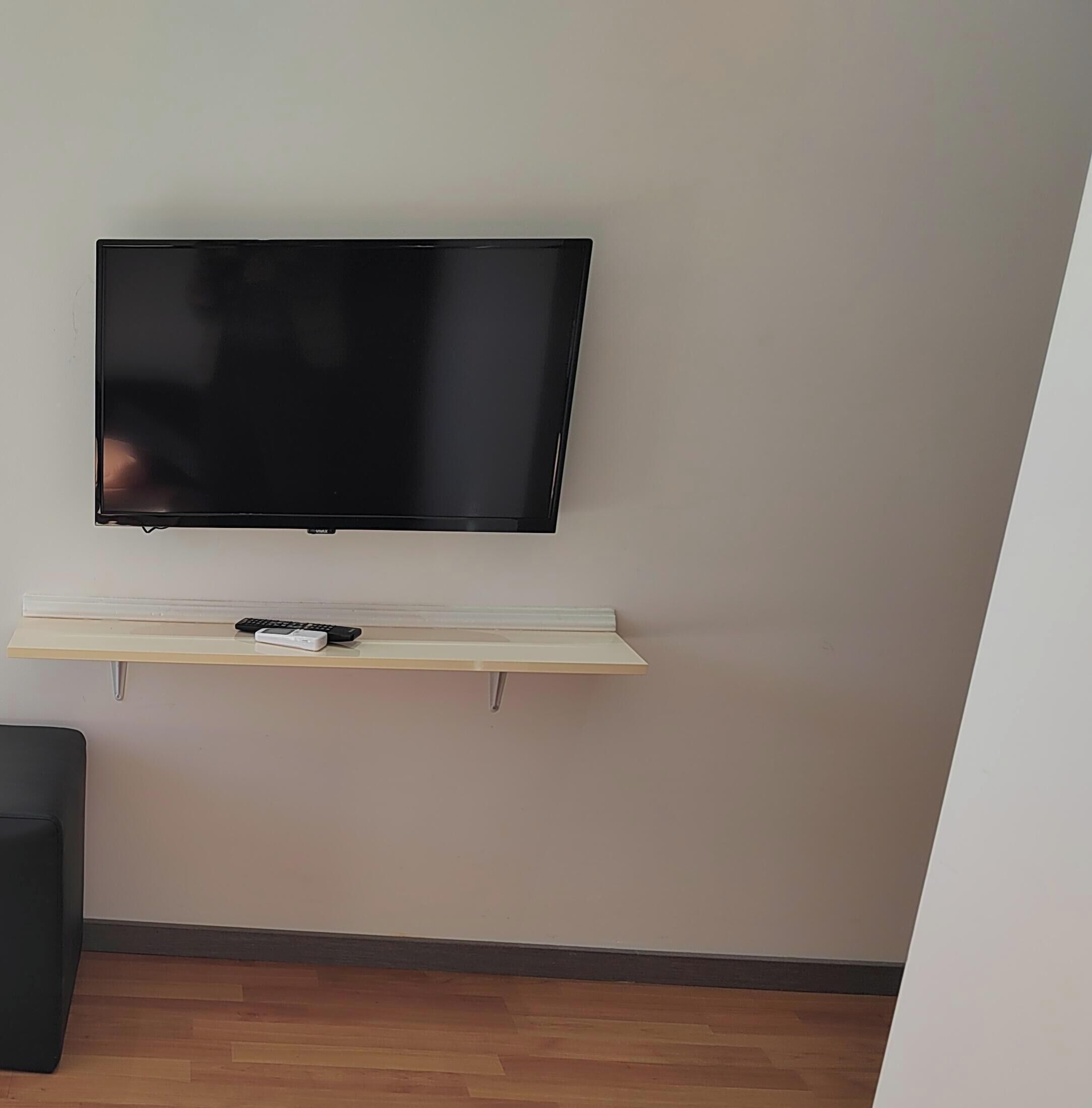 A flat-screen TV mounted on a wall above a wooden shelf with a remote control.
