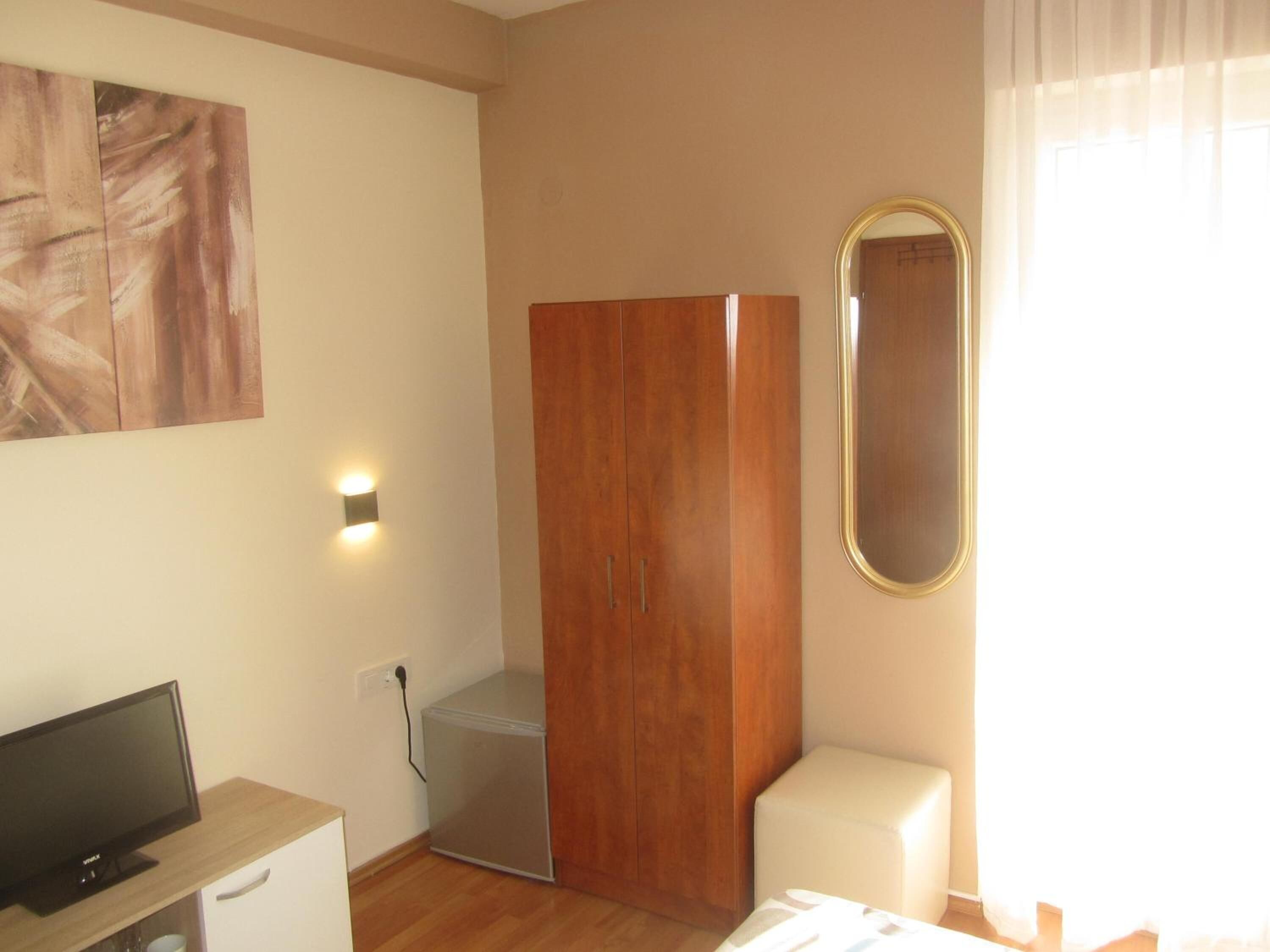 Quarto com guarda-roupa de madeira, mini-frigorífico, televisão sobre suporte de madeira e espelho na parede.