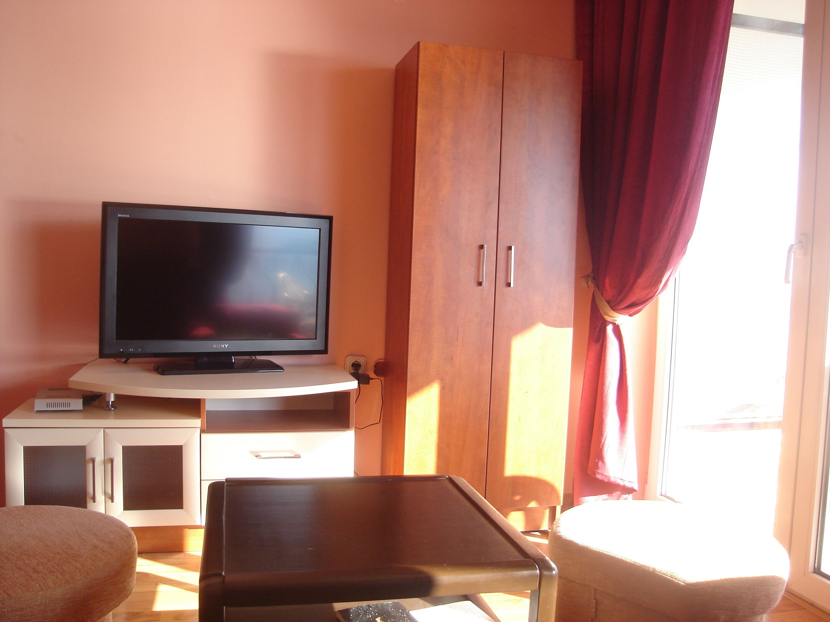 A room with a television on a stand, a wooden wardrobe, a small table, and a chair.