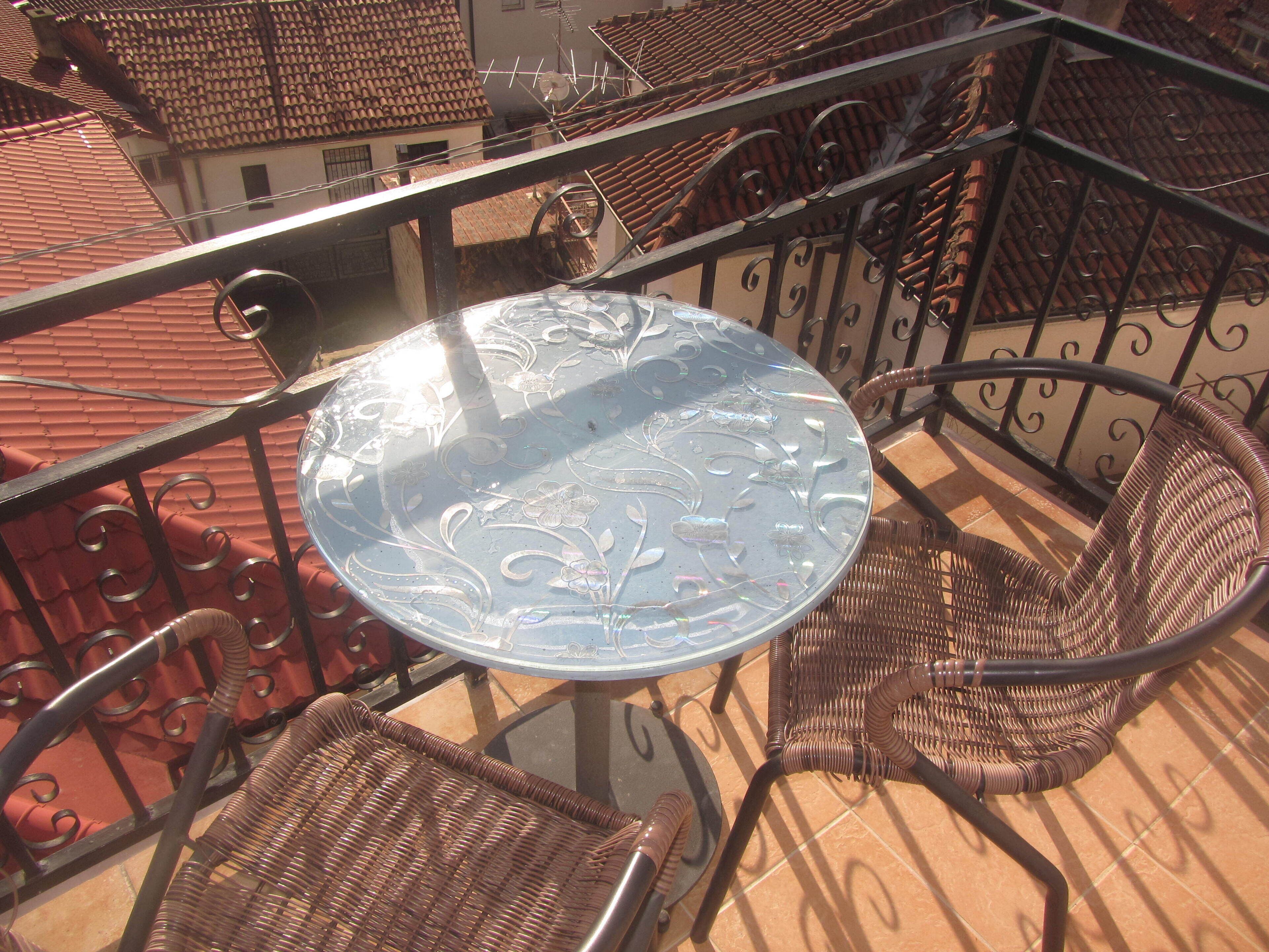 Un balcone con tavolo in vetro e sedie in vimini, con tetto in tegole e ringhiere in metallo.