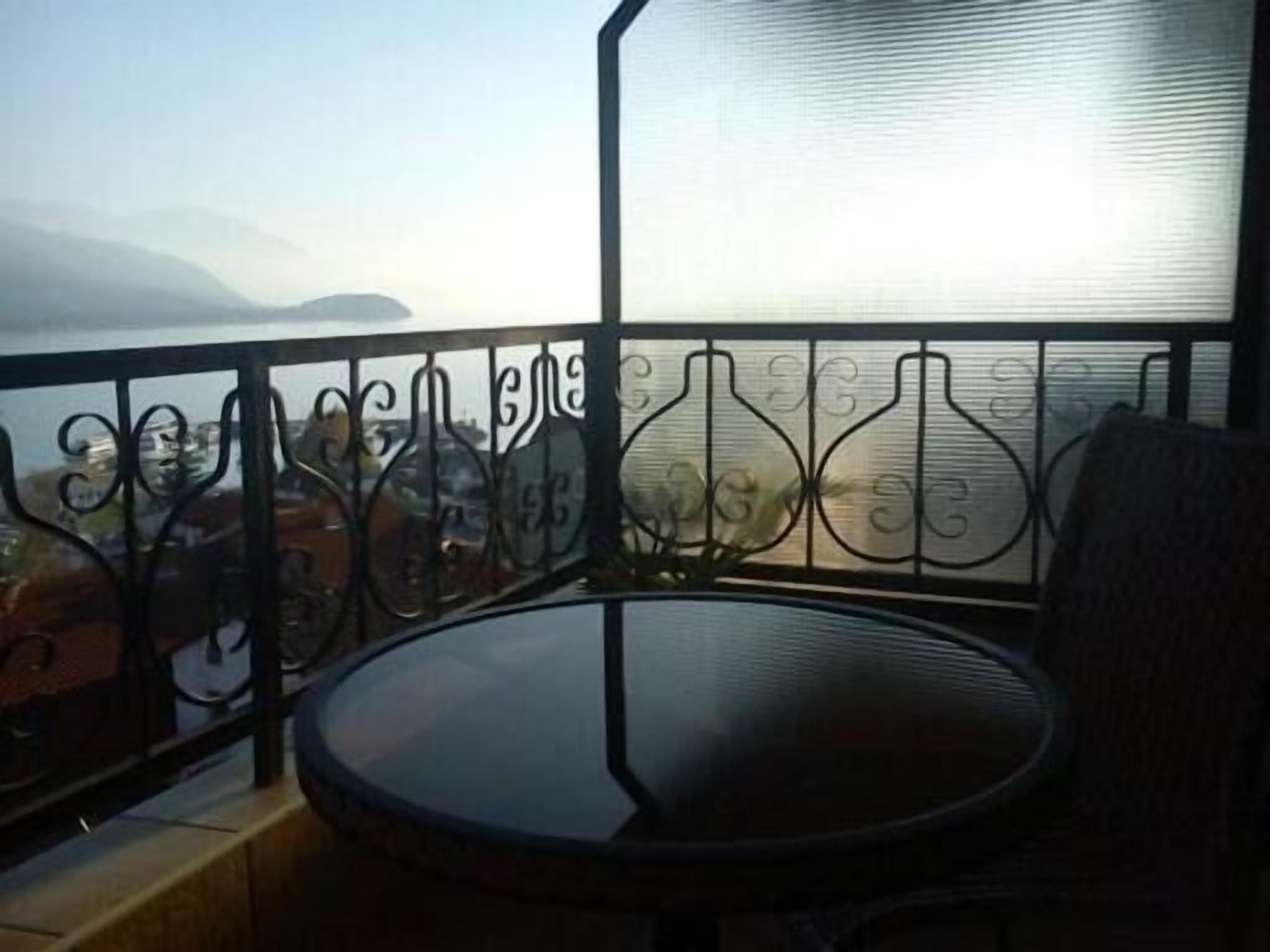 Un balcón con una mesa redonda y una silla, baranda metálica y vista al mar con montañas a lo lejos.
