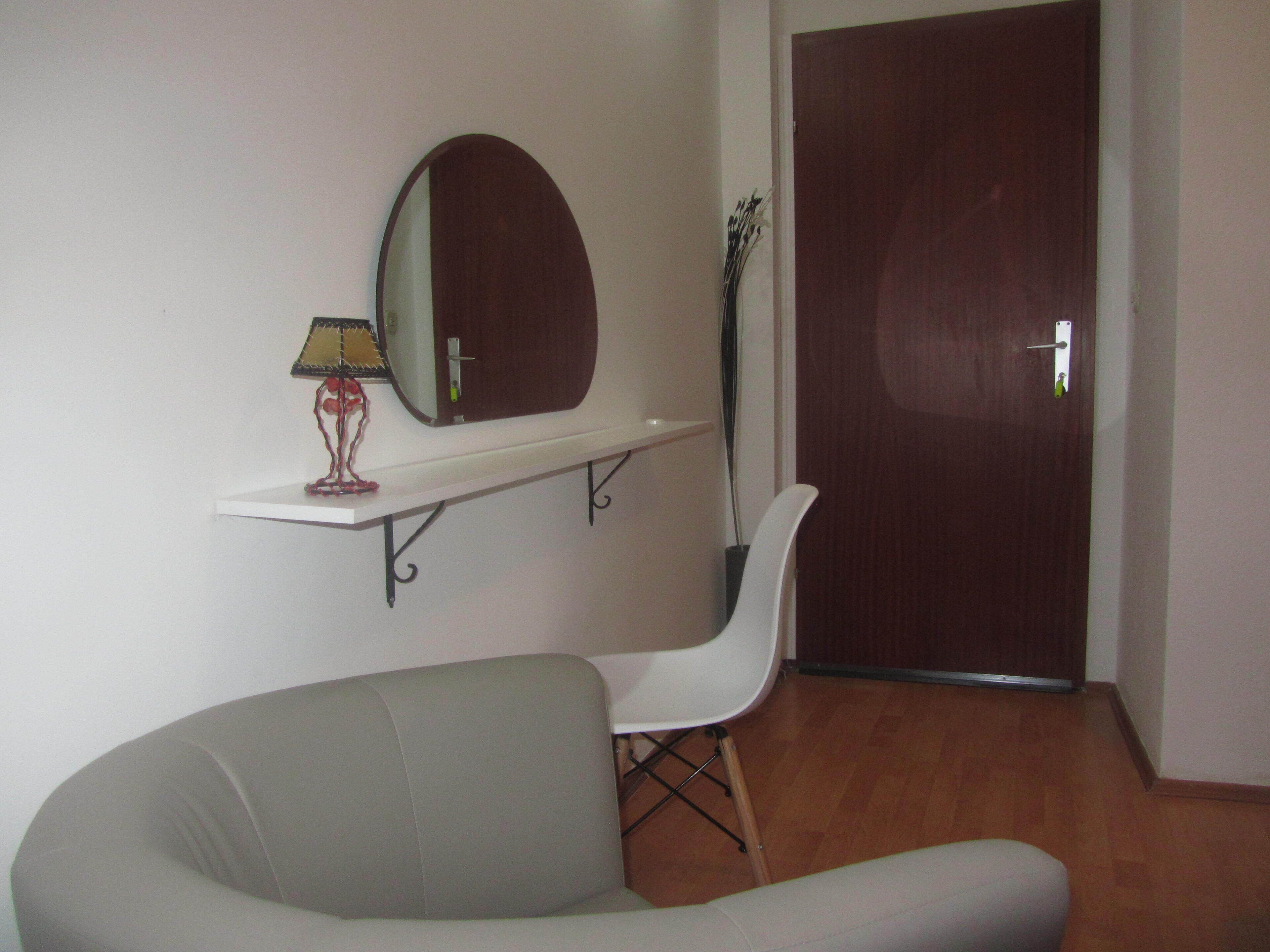 Una habitación con un sofá gris, una silla blanca, una mesita con una lámpara, un espejo y una puerta.