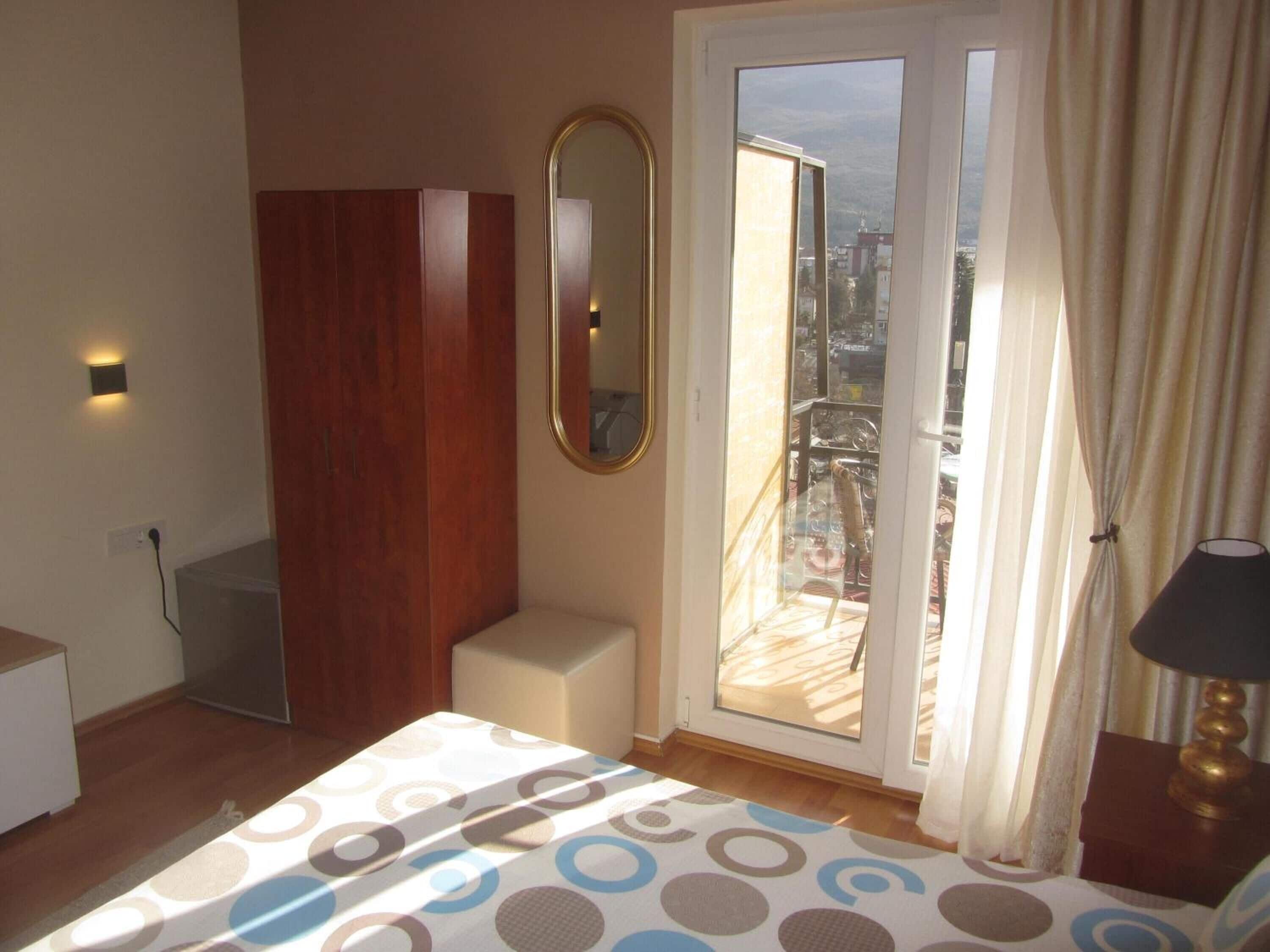 A bedroom with a bed, a wooden wardrobe, a mirror, a window with curtains, and a view of the city.