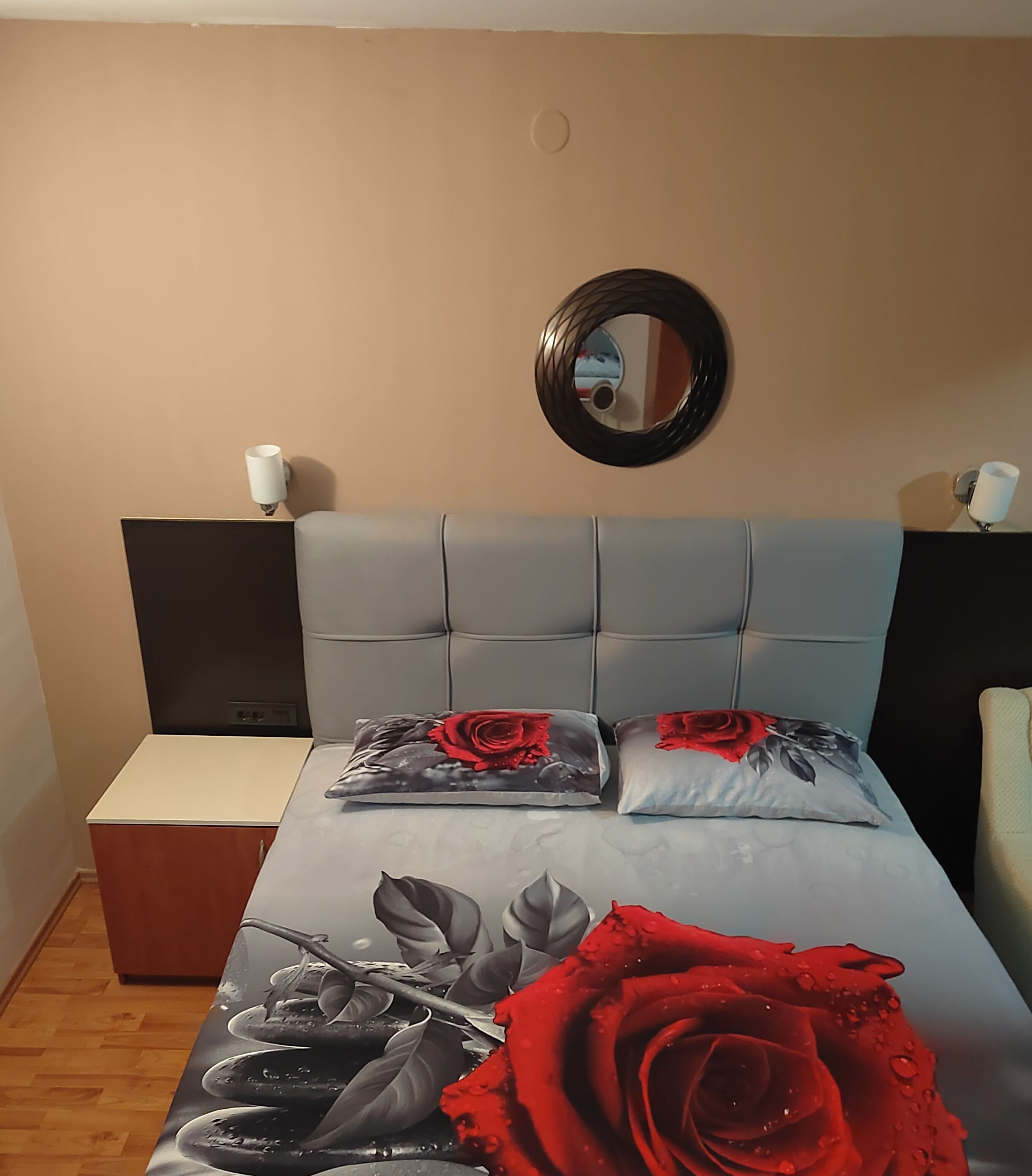 A bedroom with a grey upholstered headboard, two bedside tables, a mirror, and a bed with a floral bedspread and pillows.