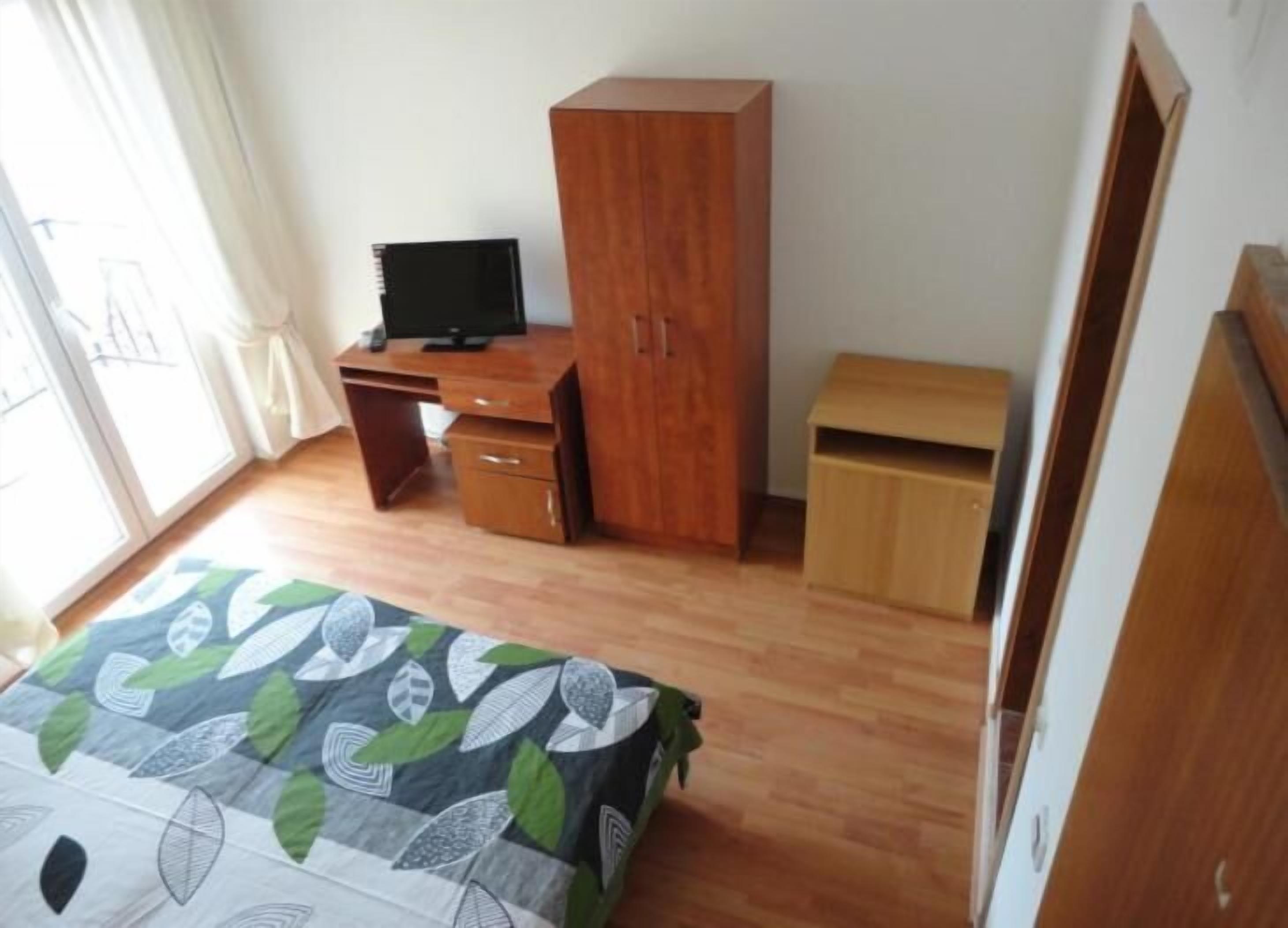 A room with a wooden desk, a television, a wardrobe, and a bed with a leaf-patterned bedspread.