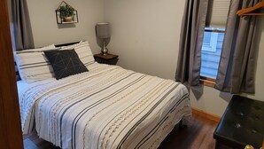 A bedroom with a bed, a bedside table with a lamp, a window with curtains, and a small plant on the wall. - Snug Harbor Inn - 2 Bedroom Cottage (Sturgeon Bay)