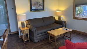 A cozy living room with a brown sofa, a wooden coffee table, and two bedside lamps. A framed artwork hangs on the wall above the sofa. - Snug Harbor Inn - 2 Bedroom Cottage (Sturgeon Bay)