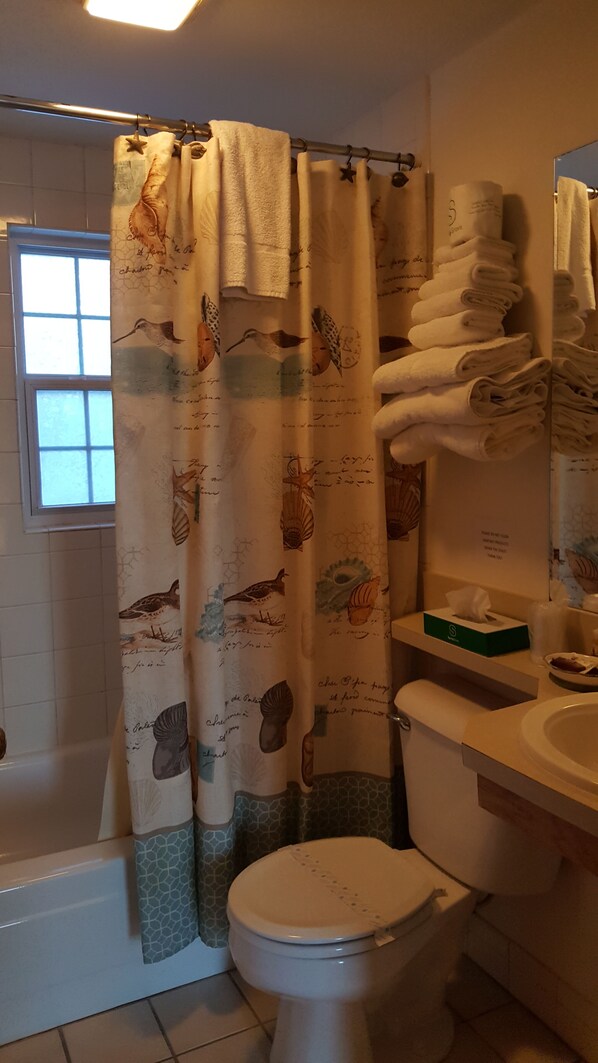 A bathroom with a shower curtain featuring bird and shell patterns, a toilet, a sink with a mirror, and a stack of folded towels.