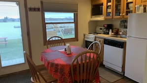 A kitchen with wooden cabinets, a stove, a microwave, and a refrigerator. There is a dining table with chairs and a window overlooking a deck. - Snug Harbor Inn - 2 Bedroom Cottage (Sturgeon Bay)
