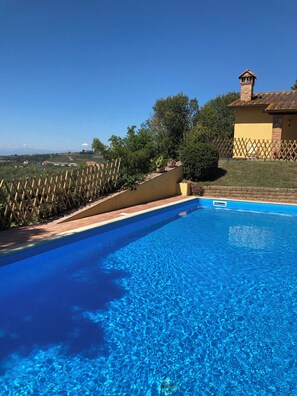 A clear blue swimming pool with a wooden fence, a yellow building with a chimney, and a view of the countryside.