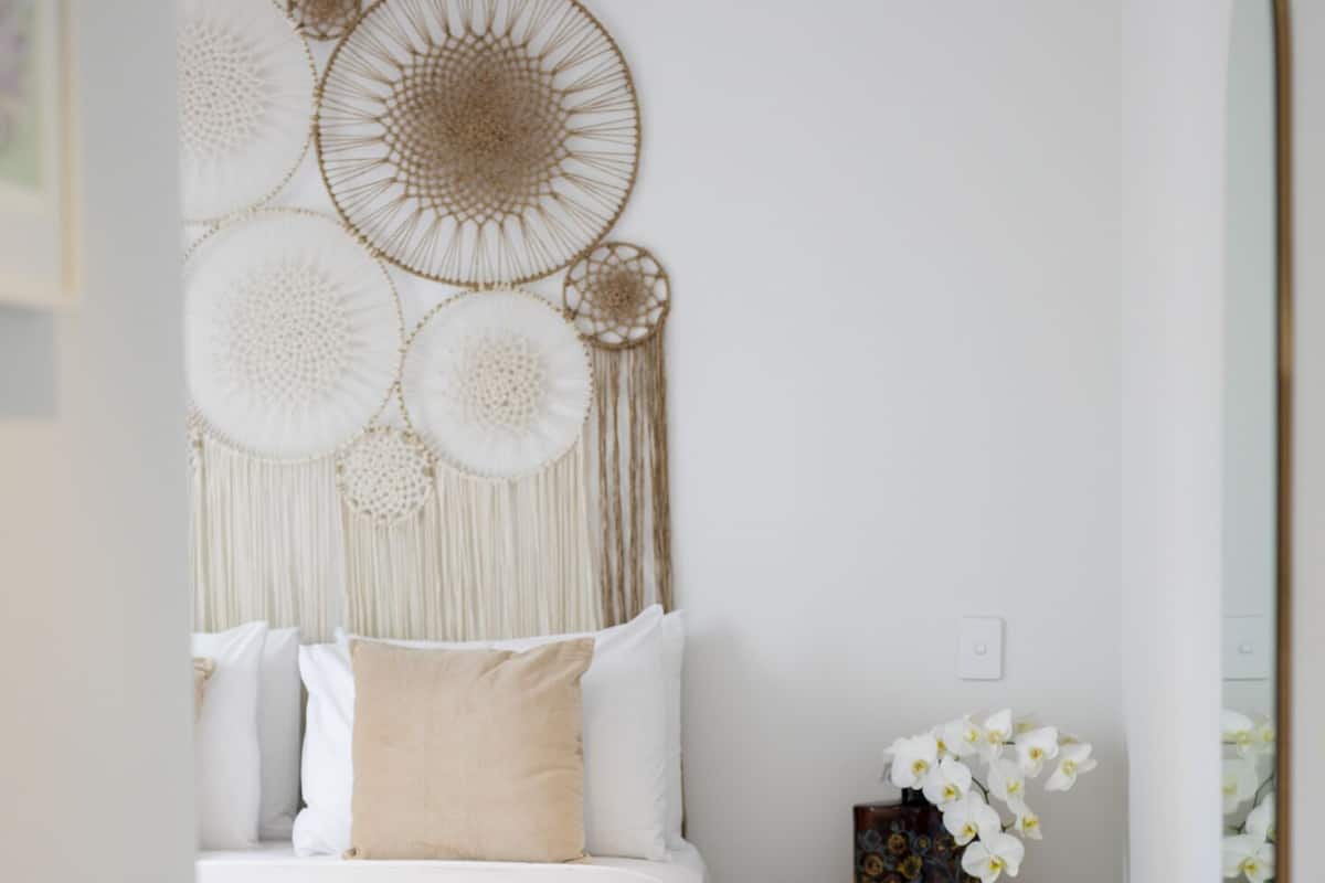 A bedroom with a bed, a bedside table with a vase of flowers, and a wall with macrame wall hangings.