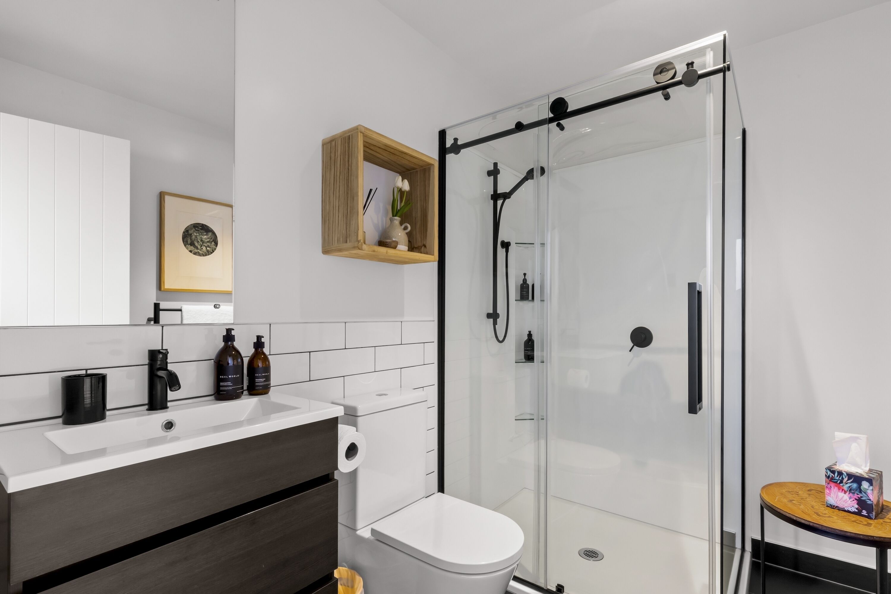 Ein modernes Badezimmer mit Duschkabine aus Glas, einer weißen Toilette, einem Waschbecken mit dunkler Arbeitsplatte und einem kleinen Holztisch mit Taschentuchbox.