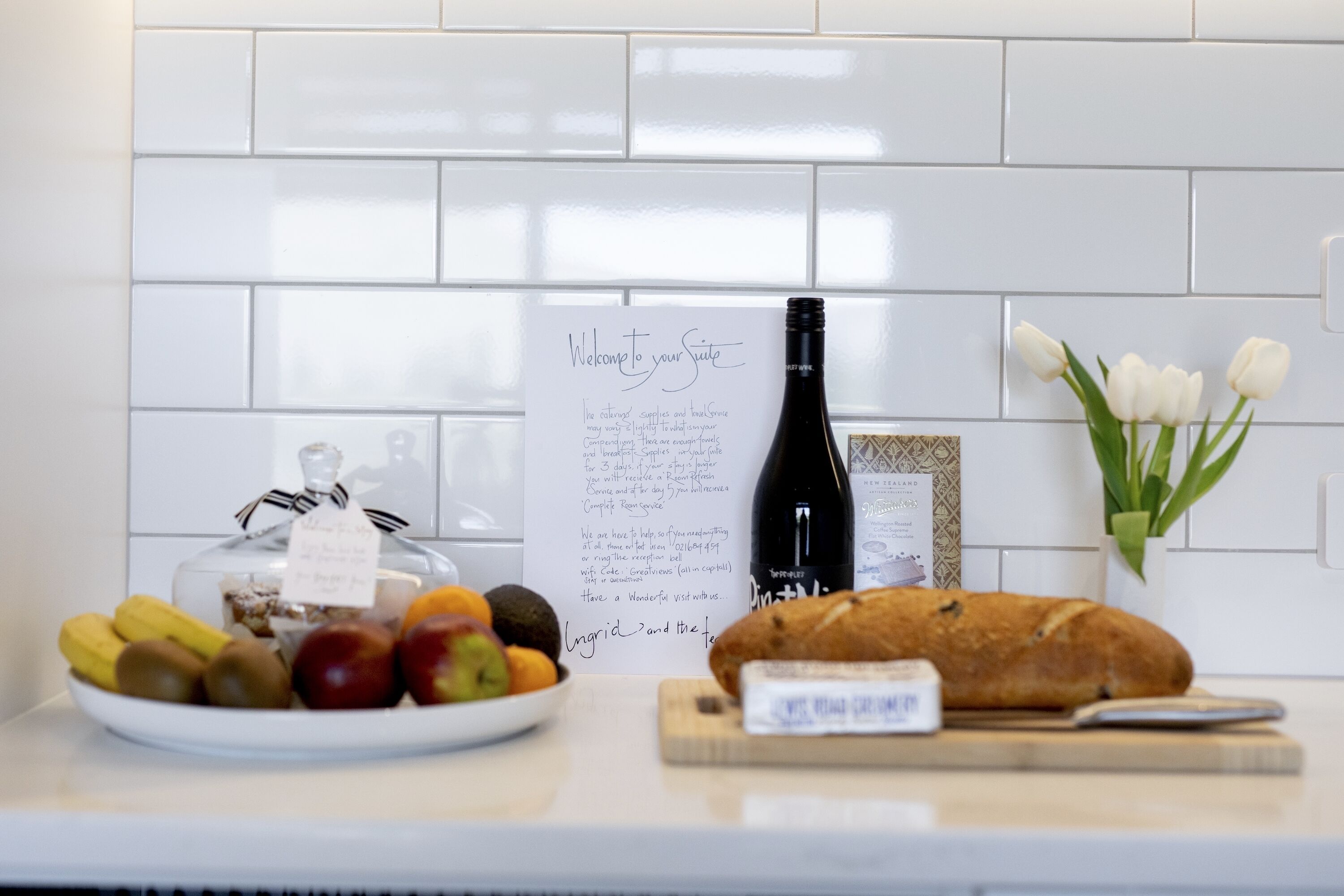 A kitchen counter with a plate of fruits, a loaf of bread, a bottle of wine, a note, and a vase with tulips.