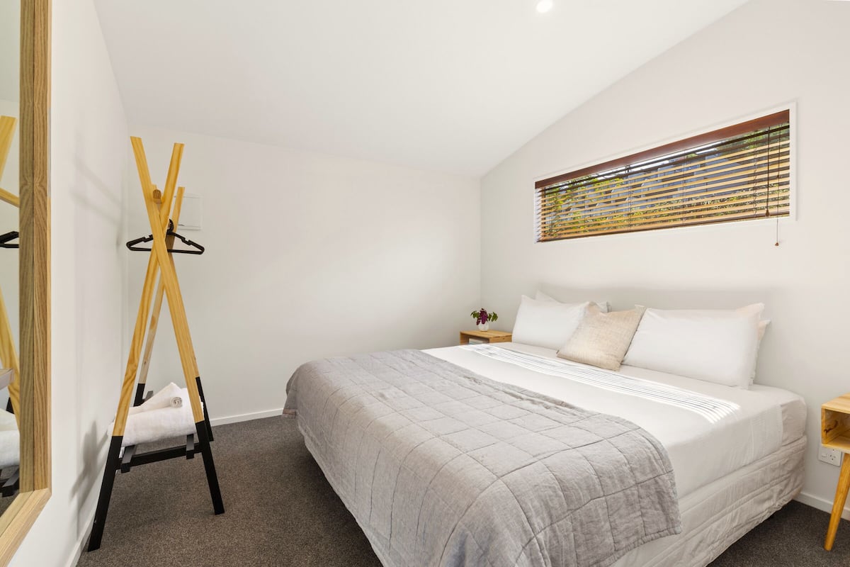 A bedroom with a bed, a wooden easel, a small table with a plant, and a window with blinds.