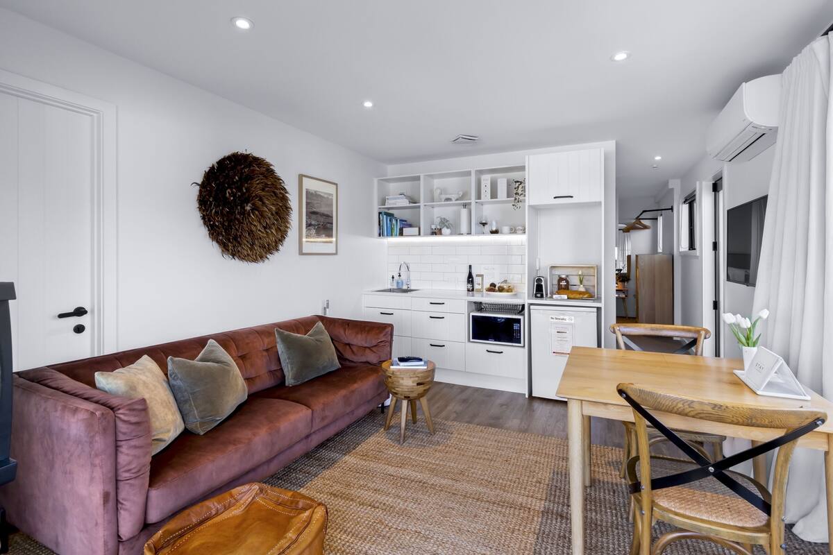 A modern living room with a brown sofa, a wooden dining table, and a kitchen area with white cabinetry.