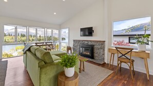 A spacious living room with a green sofa, a fireplace, a desk with a computer, and large windows offering a view of a lake and mountains. - Stay of Queenstown (Queenstown)