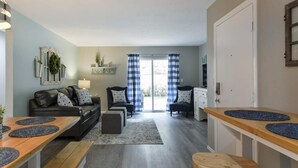 Un espace de vie compact comprenant un canapé, deux fauteuils, une table à manger avec des bancs, un coin cuisine et une porte-fenêtre coulissante donnant sur un espace extérieur.
