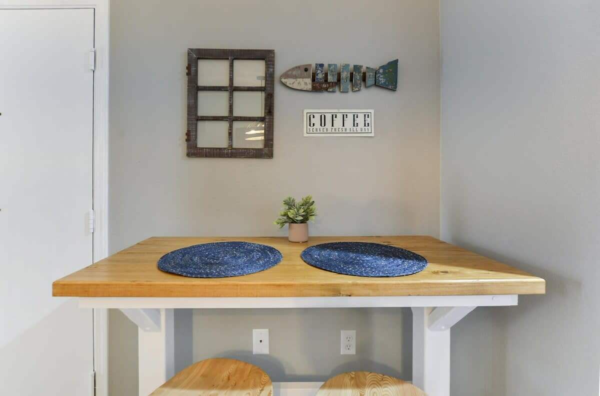Una taula de fusta petita amb dos estores blaves circulars, una planta en test, una decoració amb forma de peix, una finestra amb un dibuix de rejuntes i un cartell que diu "COFFEE".