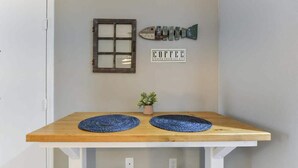 Une petite table en bois, sur laquelle reposent deux sets de table ronds bleus, un pot de fleurs, une décoration en forme de poisson, une fenêtre à carreaux et une enseigne indiquant « CAFÉ ».