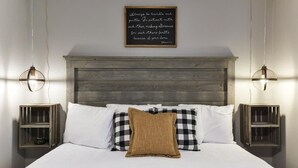 Une chambre à coucher avec une tête de lit en bois, du linge de lit blanc et des coussins à carreaux. On y trouve des tables de chevet avec des lampes et une citation encadrée au mur.