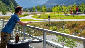 Un hombre apoyado en la barandilla de un balcón, con vista a un parque que tiene un parque infantil, una carretera sinuosa y montañas nevadas al fondo.