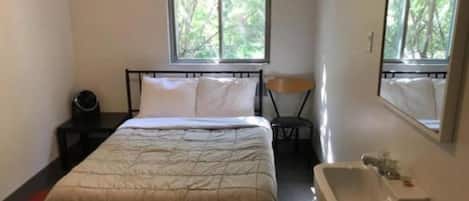 A small, simple room with a bed, a bedside table, a chair, a window, and a sink.